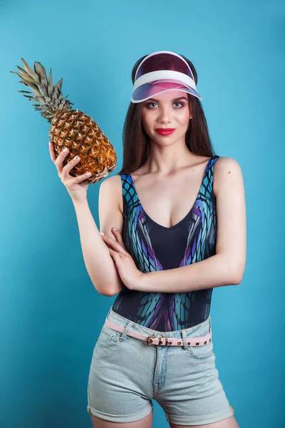 Jeune fille souriante jolie dans une visière en plastique tenant l'ananas et posant sur le fond bleu — Photo