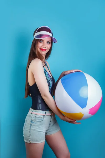 Csinos fiatal nő pózol a fürdőruhát és rózsaszín műanyag Visor sapka felfújható labdát a kék háttér — Stock Fotó