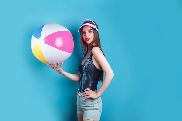 Csinos fiatal nő pózol a fürdőruhát és rózsaszín műanyag Visor sapka felfújható labdát a kék háttér — Stock Fotó