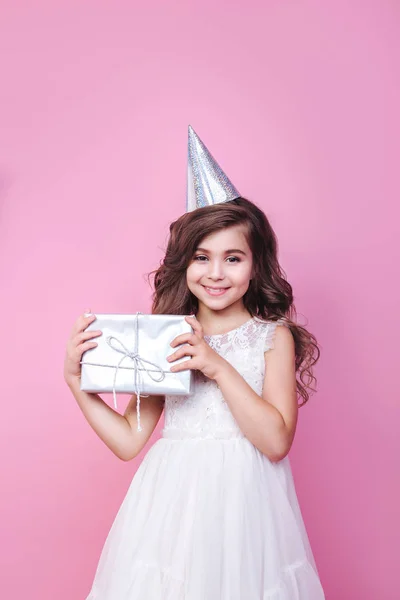 Bonita niña en vestido y gorra de cumpleaños mirando en cámara con caja de regalo en manos sobre fondo rosa — Foto de Stock