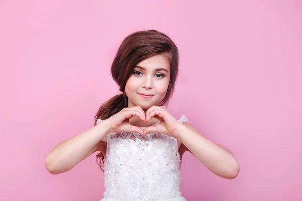 Linda niña sonriente en vestido mostrando el corazón con las manos sobre fondo rosa — Foto de Stock