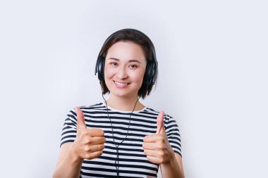 Genç Asyalı kadın siyah kulaklıklı müzik dinliyor ve baş parmağını kaldırıp el işareti yapıyor. .