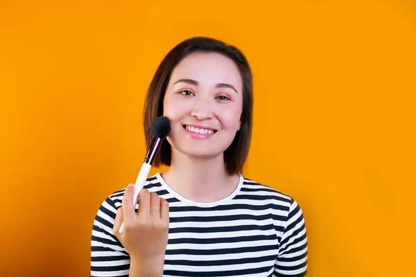 Asiática jovem bela mulher aplicando escova de pó cosmético no rosto liso isolado sobre fundo amarelo. — Fotografia de Stock
