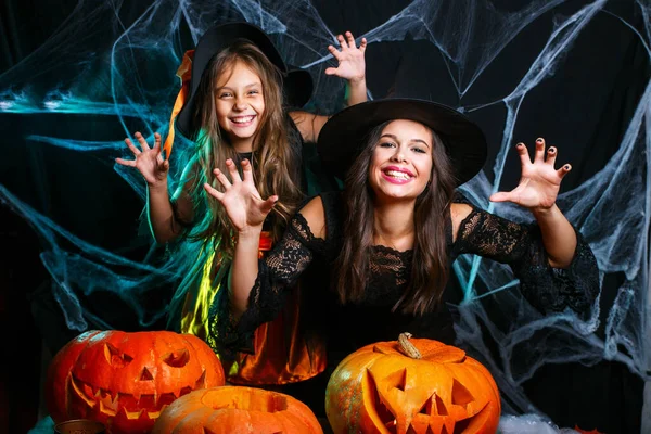 Niedliches kleines Mädchen und Mutter in Hexenhüten schauen in die Kamera und haben Spaß auf schwarzem Halloween-Hintergrund — Stockfoto