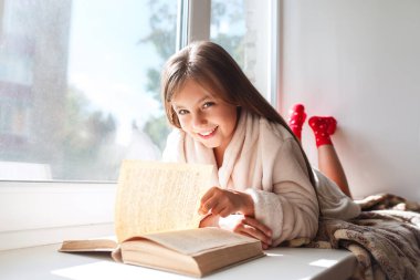Pencerenin önünde uzanmış kitap okuyan tatlı küçük bir kız.