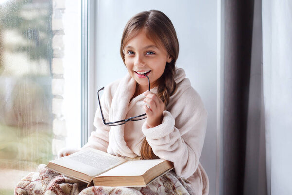 Cute child girl sitting on the window and reading a book in room at home