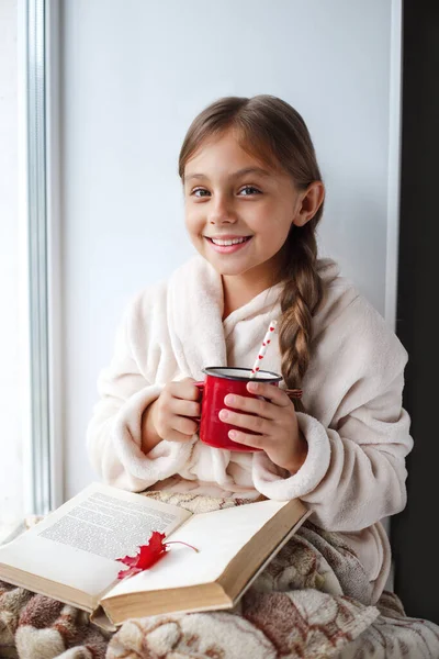 Милая маленькая девочка сидит на окне, пьет какао и читает книгу в комнате дома . — стоковое фото