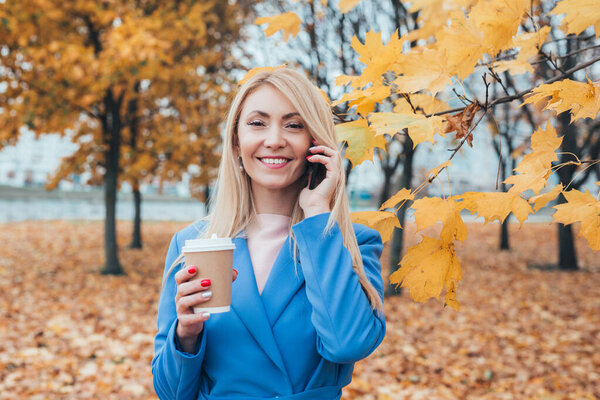 Beautiful mature woman talking on the phone and drinking coffee in autumn park