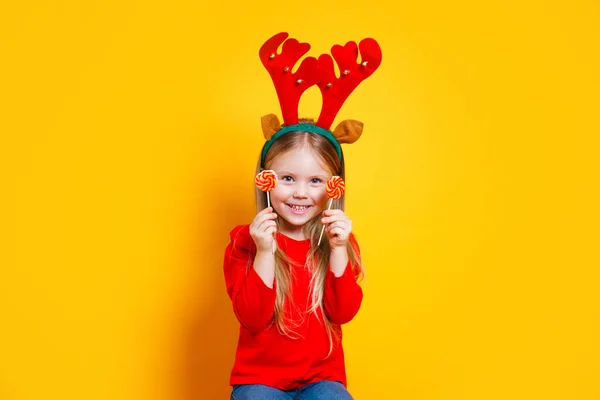 Feliz niña divertida en traje de renos de Navidad rojo sosteniendo piruleta sobre fondo amarillo —  Fotos de Stock