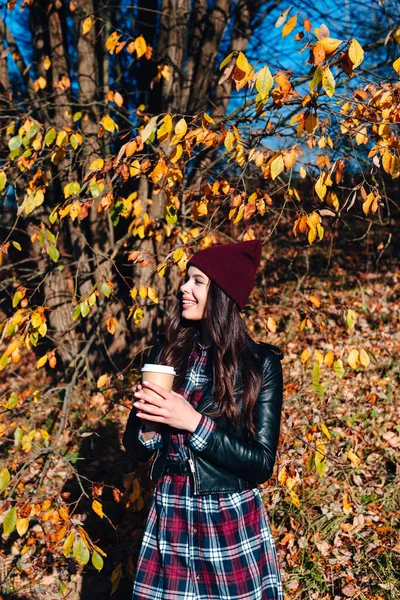 Stylish young woman in a hat and a plaid shirt drinking coffee in autumn park — Stock Photo, Image