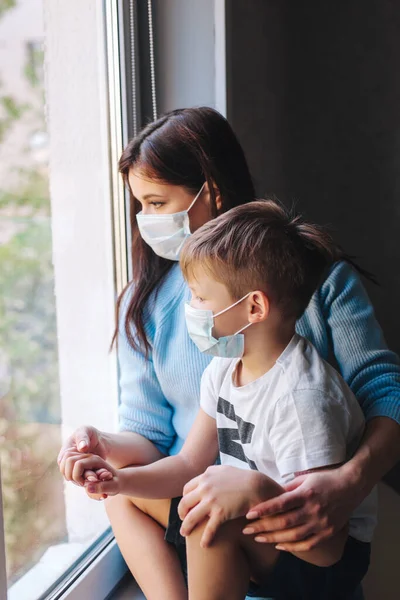 Madre y su hijo con máscaras protectoras muy tristes mirando por la ventana. Covid-19 pandemia y cuarentena — Foto de Stock