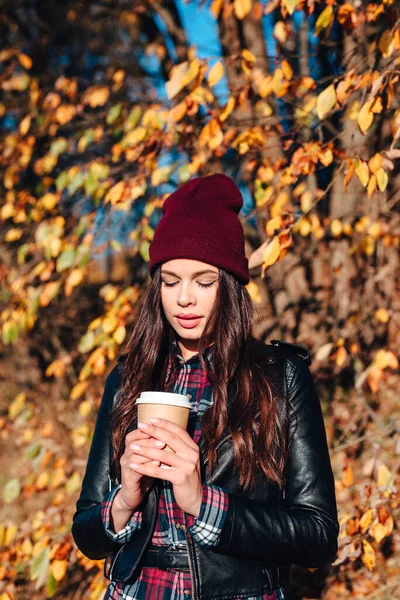 Stilvolle junge Frau mit Hut und kariertem Hemd trinkt Kaffee im Herbstpark — Stockfoto