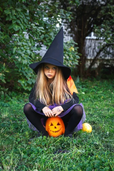 Menina engraçada em traje de bruxa para o Halloween sentado na abóbora Jack ao ar livre — Fotografia de Stock