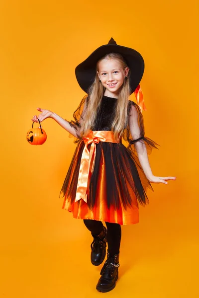 Bonito menina feliz vestida como bruxa com balde de doces no fundo laranja — Fotografia de Stock