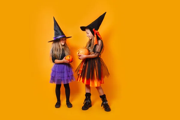 Δύο κοριτσάκια με αποκριάτικες στολές μαγισσών διασκεδάζουν σε πορτοκαλί φόντο. — Φωτογραφία Αρχείου