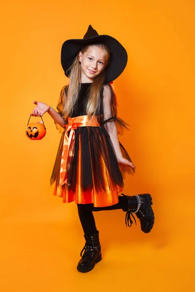 Bonito menina feliz vestida como bruxa com balde de doces no fundo laranja — Fotografia de Stock