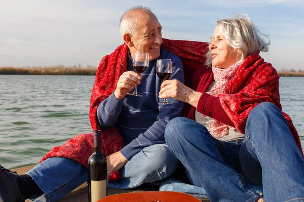 Portrait of happy senior couple drinking red wine by the lake on sunny autumn day.