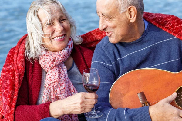 Portrait of happy senior couple drinking red wine and playing guiar by the lake on sunny autumn day.
