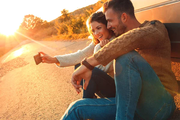Beautiful couple on road trip, they are taking a break from driving leaning against a old fashioned car and taking selfie