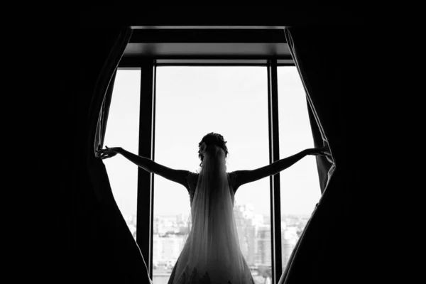 ウエディングドレスとベールの若い花嫁のバックビューは 窓と開口部のカーテンの近くに立って 黒と白の画像 — ストック写真