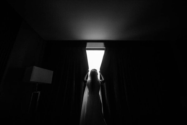 back view of young bride in wedding dress and veil opening curtains, black and white image
