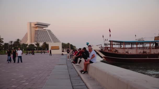 日落时的多哈小巷 人们坐着散步 背景是悬挂卡塔尔国旗的独桅帆船和喜来登酒店 — 图库视频影像