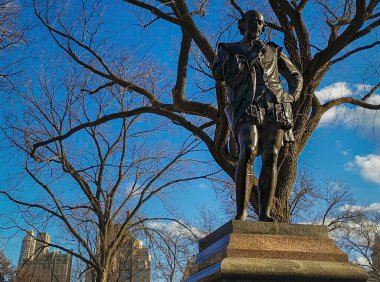 William Shakespeare 'in heykeli (John Quincy Adams Ward) Central Park' ta duruyor. New York şehir gündüz manzaralı ağaçlar, gökdelenler ve gökyüzünde bulutlar var.