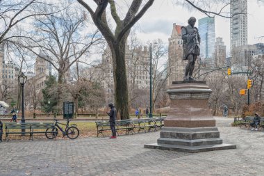 William Shakespeare 'in heykeli (John Quincy Adams Ward) Central Park' ta duruyor. New York şehir gündüz manzaralı ağaçlar, gökdelenler ve gökyüzünde bulutlar var.
