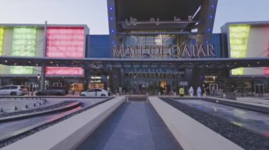 Doha 'daki Katar alışveriş merkezinin ana girişinde günbatımında güvenlik kameraları, alışveriş merkezine giren insanlar ve arabalar görülüyor.