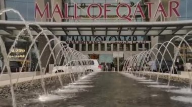 Gün batımında Doha Katar 'daki Katar' daki Mall of Katar 'ın ön plandaki çeşmeye yakınlaşması, arka plandaki ana giriş kapısı, insanların ve arabaların hareket etmesi.