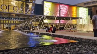 Gece Doha Katar 'daki Katar Alışveriş Merkezi' nin hızlandırılmış görüntüsü ön planda fıskiyeler ve arka planda insanlar ve arabaların hareket ettiği ana giriş kapısı olan panoramik bir manzara gösteriyor.