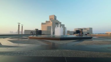 Doha Katar 'daki İslam Sanatları Müzesi ön planda fıskiyeyi ve arka planda müzenin ana girişini gösteren otomobil parkı ve yürüyen insanları gösteren bir çekim yapıyor.