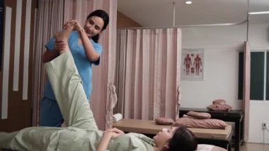 İspanyol kadın fizyoterapist Asyalı hastanın bacaklarını masaj yatağında geriyor, sağlık tedavisi görüyor, diz kası ağrısı tedavisi görüyor ve sağlık merkezinde pasif tıbbi tedavi uyguluyor..