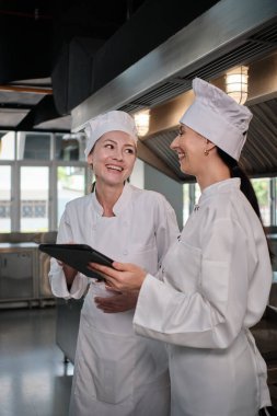 Önlüklü iki beyaz kadın gurme aşçı malzeme hazırlığı ve tablet üzerine menü, mutfakta yemek pişirme, lezzetli mutfaklarda, yemek restoranlarında ve mutfak yemeklerinde profesyonel partnerler hakkında konuşuyor..