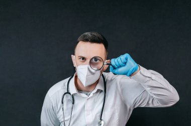 Tıbbi maskeli ve mavi eldivenli olgun bir erkek doktor koyu arka planda duruyor..