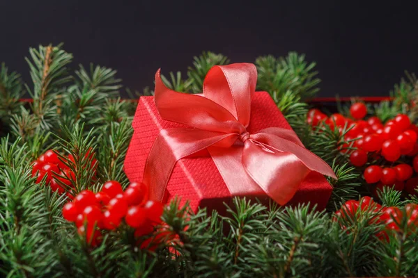 Roter Geschenkkarton mit satinrosa Schleife, eingetaucht in die Nadeln eines mit roten Beeren geschmückten Weihnachtsbaums. — Stockfoto