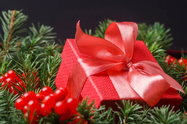 Červená dárková krabička s saténovou růžovou mašlí, ponořená do jehličí vánočního stromku zdobeného červenými bobulemi. — Stock fotografie