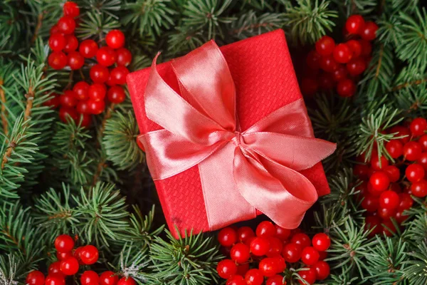 Caja regalo regalo rojo con lazo rosa satinado, inmerso en las agujas de un árbol de Navidad decorado con bayas rojas . —  Fotos de Stock