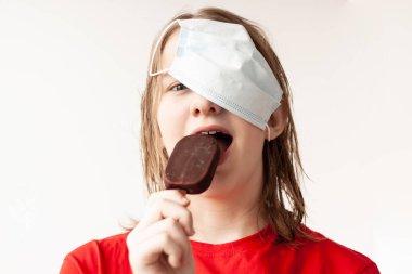 Sarı saçlı genç bir kız saçına tıbbi bir maske takmış, çikolatalı dondurmayı ısırmış, seçici bir odak noktası var..