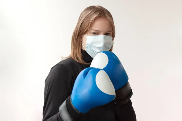 A girl with blond hair in a medical mask and blue Boxing gloves stands in a boxer\'s pose, right hand in front of the left near the face.
