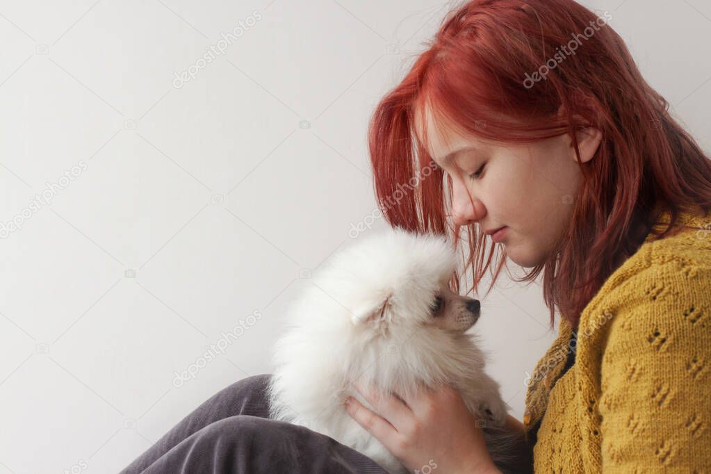 A teenage girl with bright red hair in a yellow, knitted jacket and gray trousers, in profile, holds a white fluffy Pomeranian puppy in front of her face