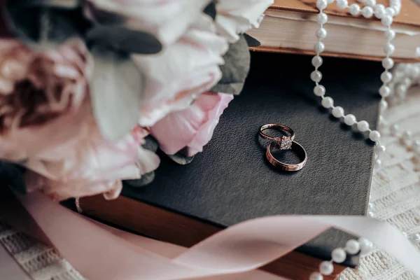 Свадебные кольца лежат на черной книге возле свадебного букета, атласных лет и белых бусин — стоковое фото