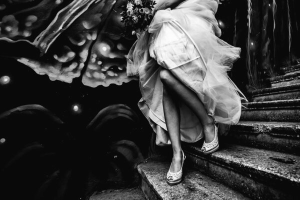 Bride in white shoes runs on a concrete staircase with graffiti, holding a dress and a beautiful wedding bouquet in black and white