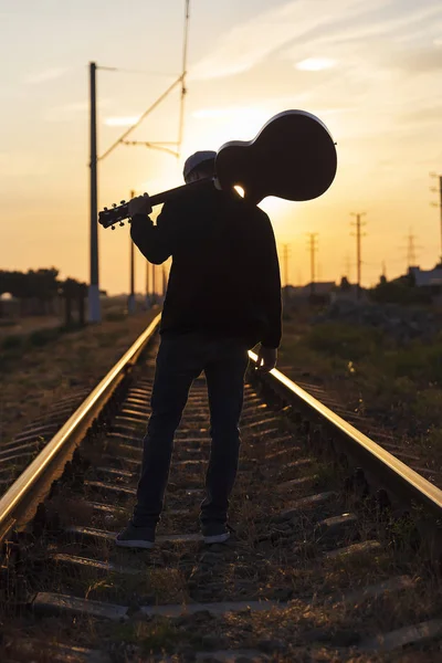 Een jonge kerel staat op de rails met een gitaar op zijn schouders een — Stockfoto