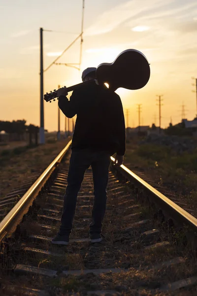 Genç bir adam omuzlarında bir gitar ile raylar üzerinde duruyor