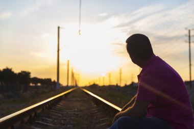 Genç bir adam rayların üzerinde oturur ve güneşte mesafeye bakar