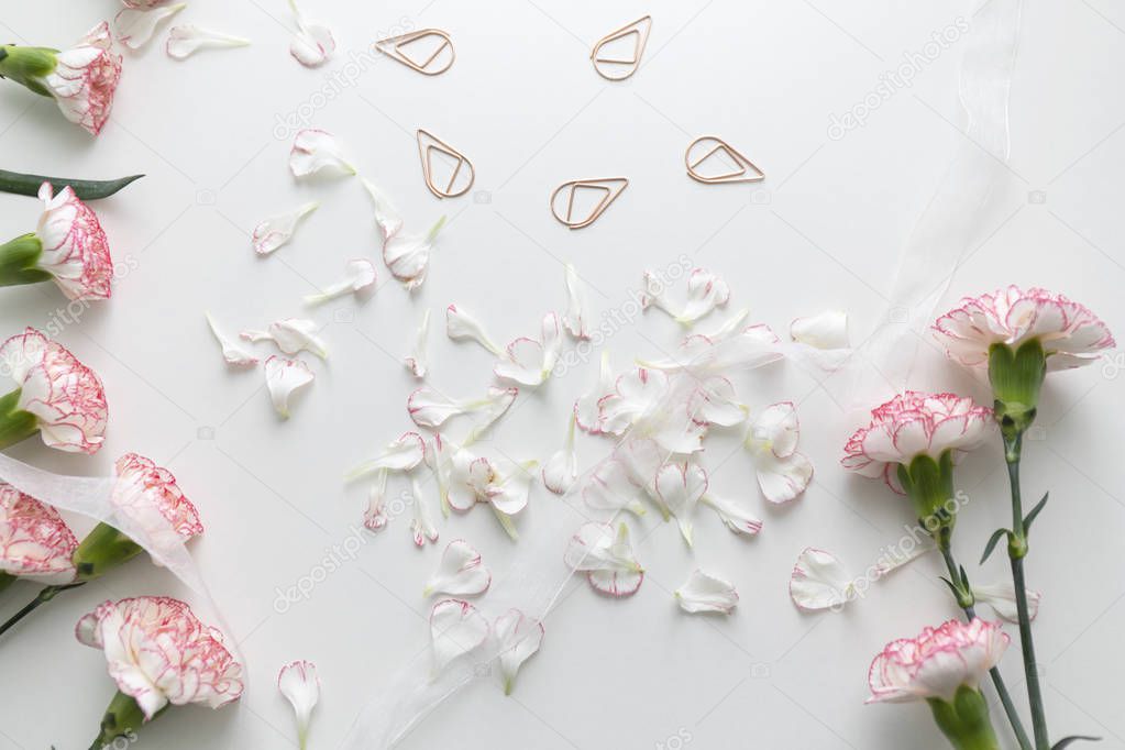 Flat lay of pink and white carnation flowers and petals with white ribbon and binder clips on white desk.