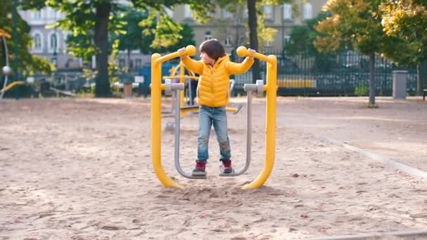 Um menino de roupas amarelas está fazendo um exercício no simulador amarelo . — Vídeo de Stock