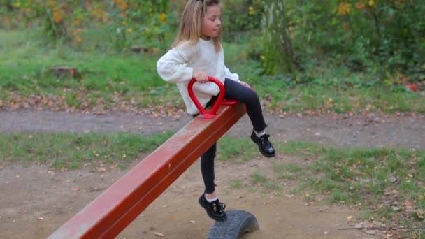 Uma menina de roupas brancas está montando um balanço de equilíbrio em um parque da cidade — Vídeo de Stock