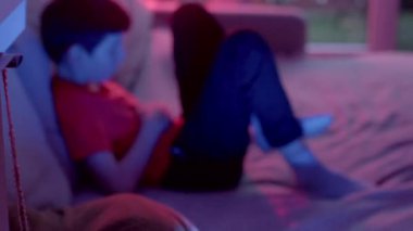 A young boy lounging on a couch, immersed in a digital device, illuminated by vibrant neon lights, creating a cozy atmosphere.
